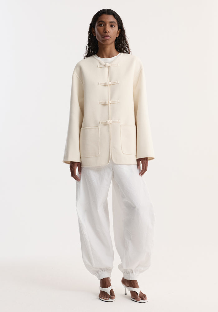 Woman wearing double-faced wool pankou jacket with fabric toggle closures, embroidered edges, and patch pockets, paired with white drawstring trousers and heeled sandals.