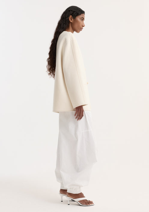 Woman wearing double-faced wool pankou jacket with fabric toggle closures, embroidered edges, and patch pockets, paired with white drawstring trousers and heeled sandals.