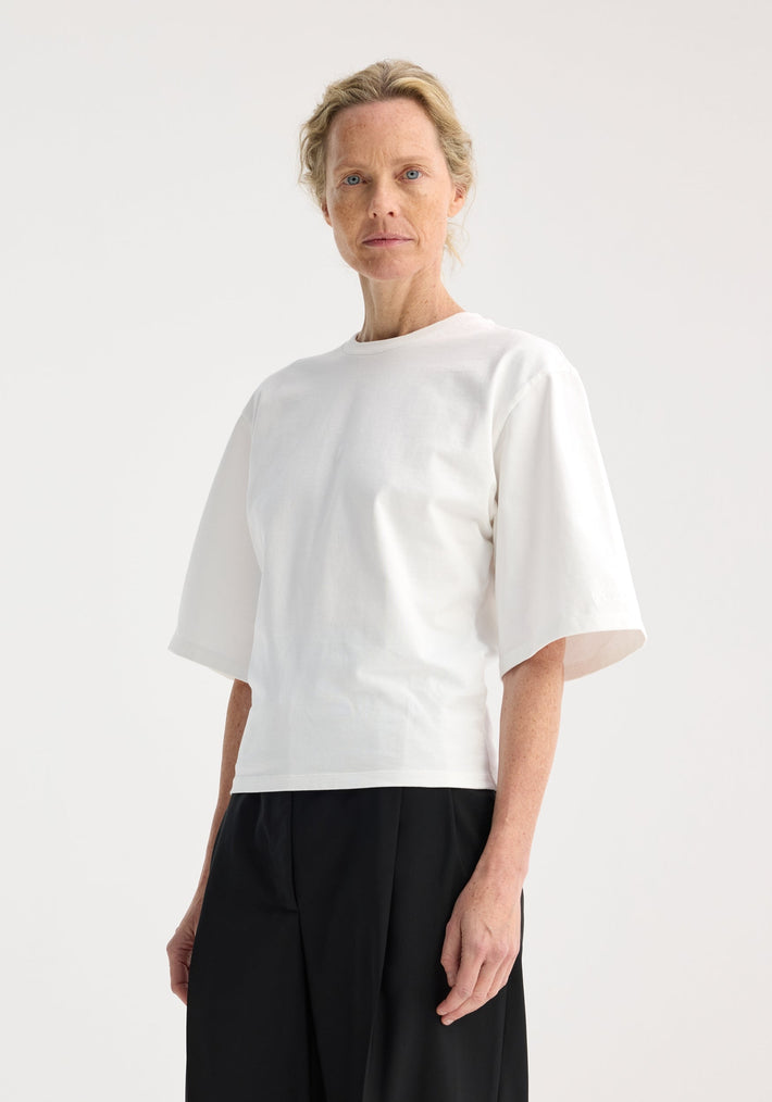 Person wearing Fitted logo t-shirt in optic white with a crew neck, tapered elbow-length sleeves, and logo embroidery, paired with loose black trousers.