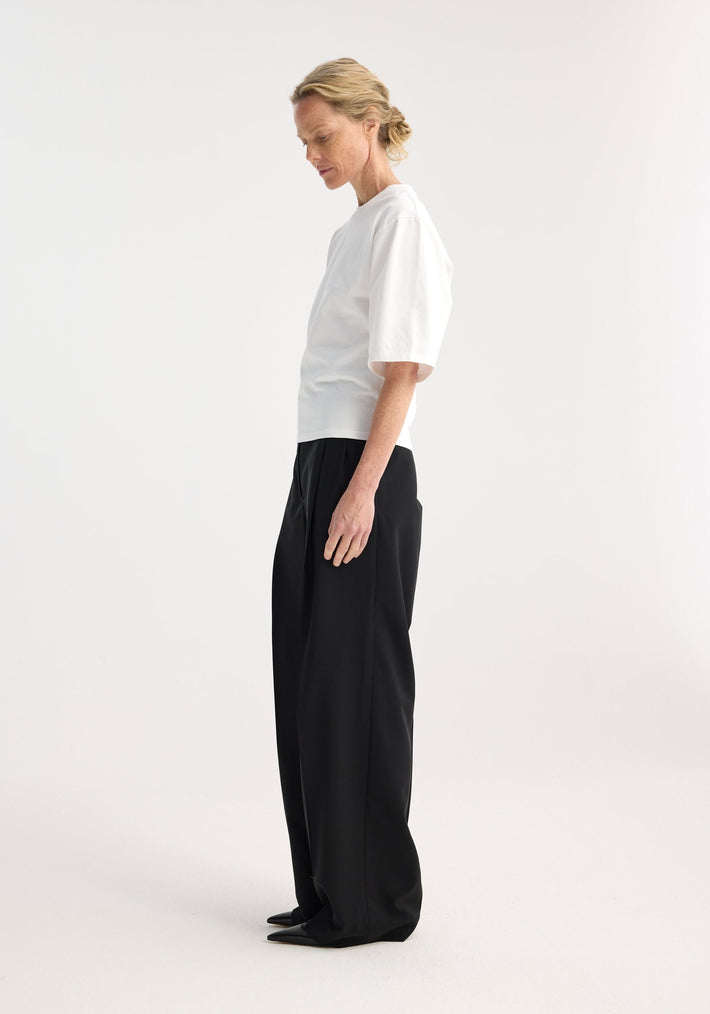 Fitted logo t-shirt in optic white, worn by model with elbow-length sleeves, crew neck, and subtle logo on left sleeve, paired with wide-leg trousers.