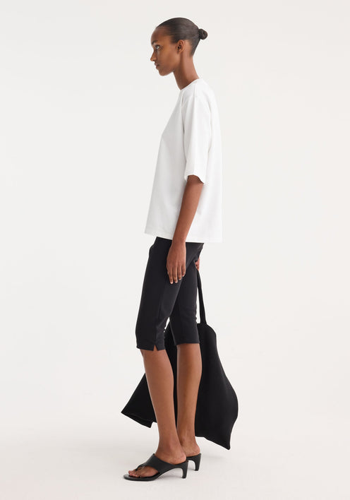 Woman wearing Scuba capri | black, showcasing tailored, knee-length scuba fabric trousers with a structured fit, styled with a loose white T-shirt and black sandals.