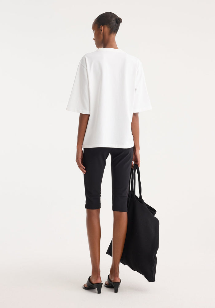 Scuba capri | black: Model in tailored, capri-length black scuba leggings, holding a slouchy black tote, styled with a white T-shirt and open-toe heeled mules.