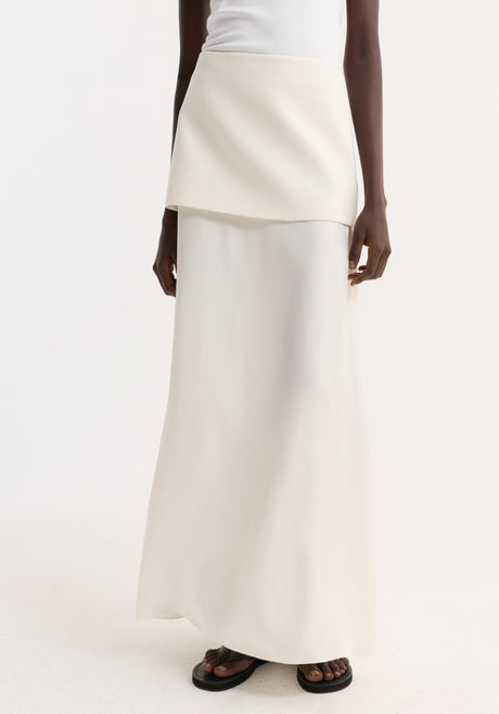 Model wearing Integrated fluid silk skirt, cream, with asymmetrical overlay and high waist, paired with white tank, black bucket hat, and black sandals.