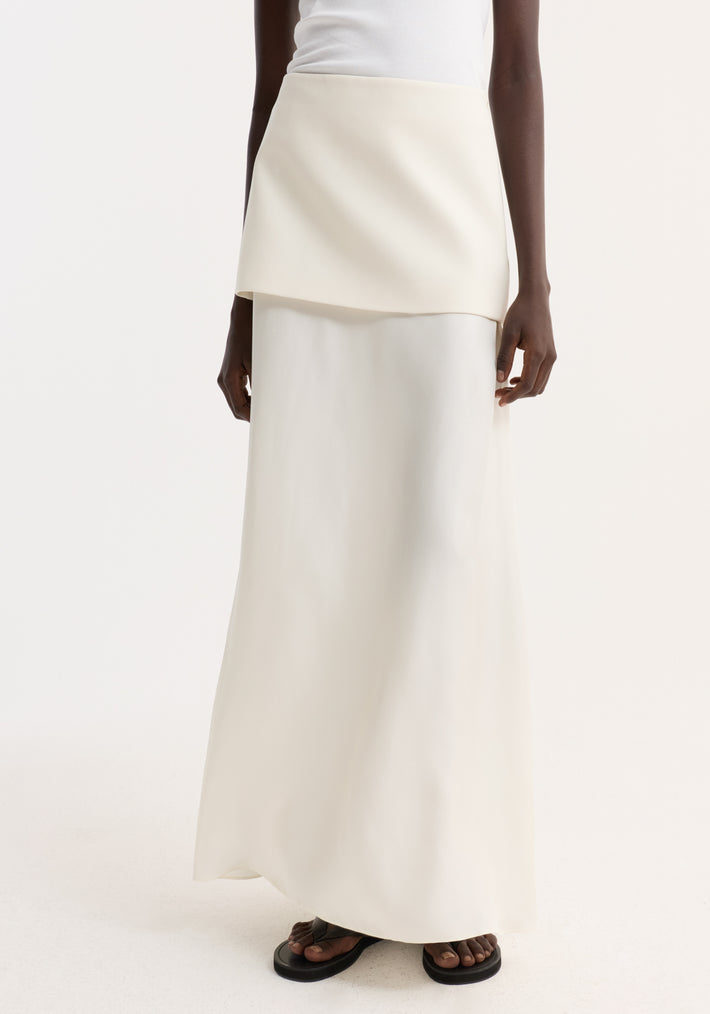 Integrated fluid silk skirt in cream, featuring a high-waisted, floor-length design with a sculptural silhouette, worn with black sandals against a neutral backdrop.