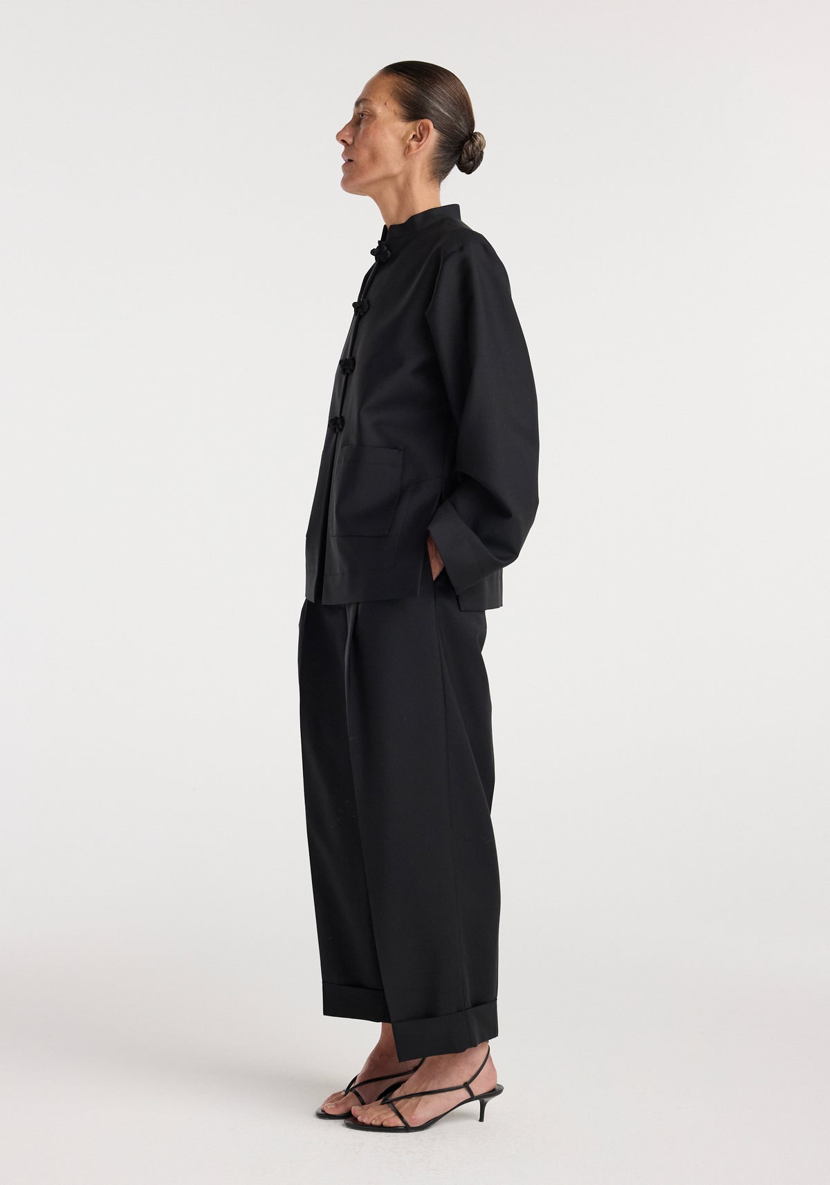 Side view of a woman wearing a black Pankou closure jacket | black with long sleeves and large front pockets, paired with wide-leg black pants and black strappy heels.