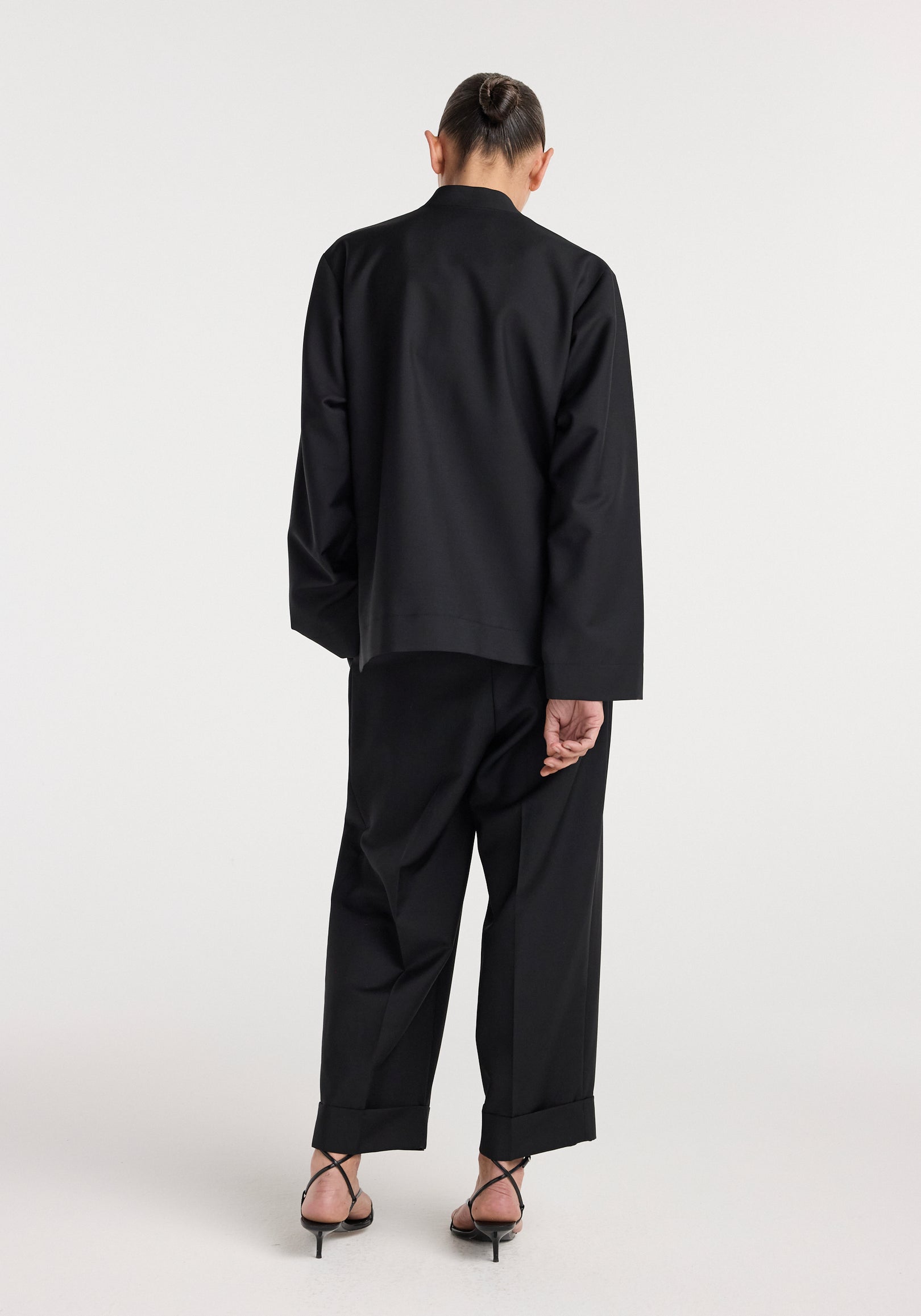 Rear view of a person wearing a black Pankou closure jacket | black with matching black wide-leg pants and strappy black heels against a plain background.