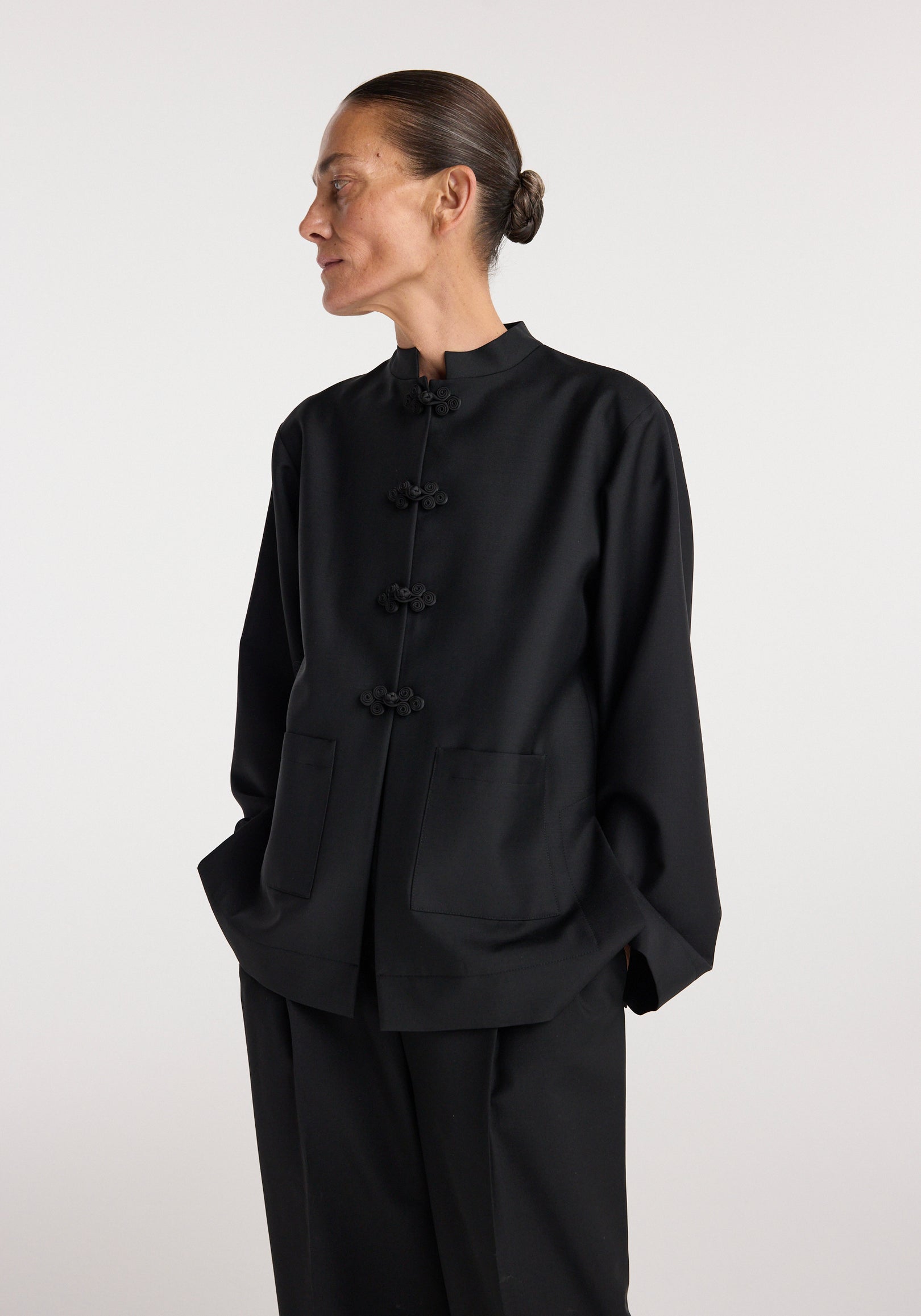Woman wearing a black Pankou closure jacket | black with traditional knot buttons, large front pockets, and a mandarin collar, styled with matching black pants.