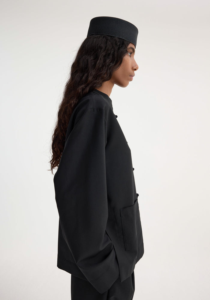 Side-profile of a woman wearing the Mandarin jacket | black, featuring a collarless design, pankou closures, and patch pockets, paired with matching trousers and a pillbox hat.