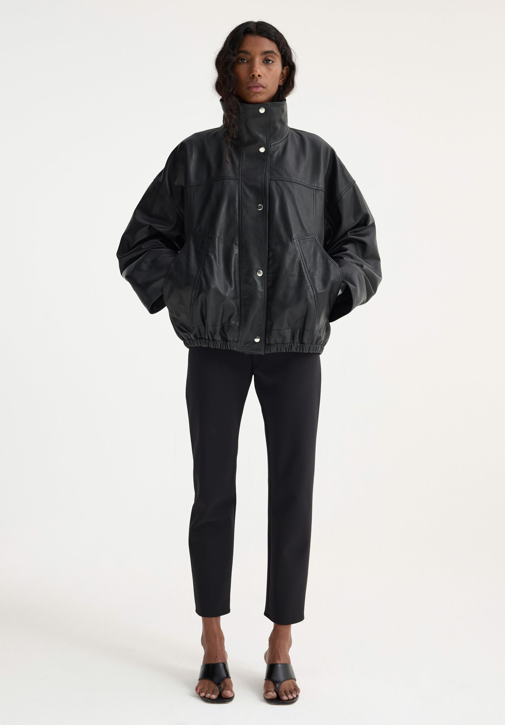 Model wearing a black oversized leather jacket with snap buttons and elastic hem, paired with slim black pants and black open-toe sandals, showcasing the Reinvented leather jacket | black.