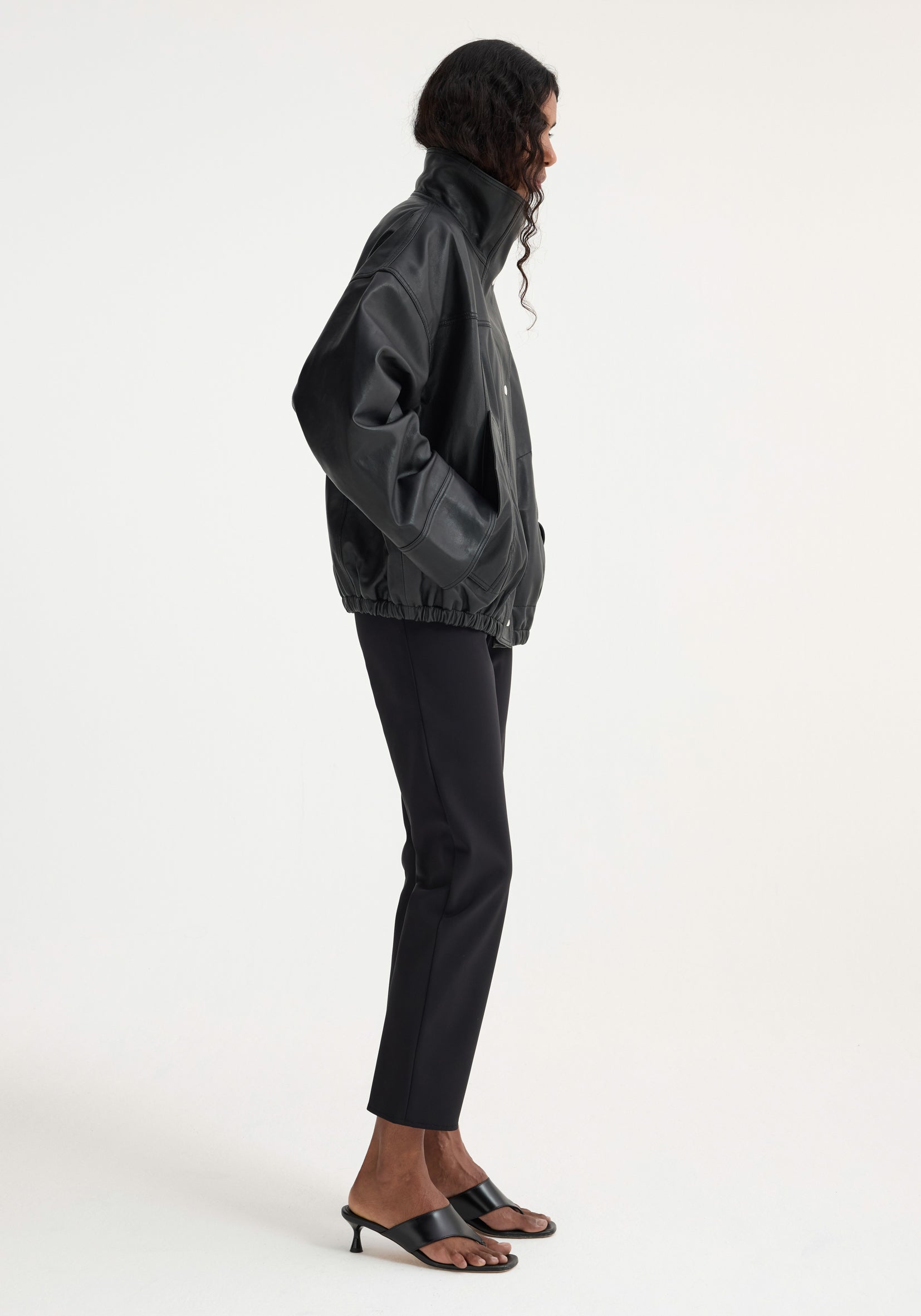 Side view of a woman wearing a high-collared black soft leather jacket with elastic hem and long sleeves, paired with slim black pants and black heeled sandals, Reinvented leather jacket | black.