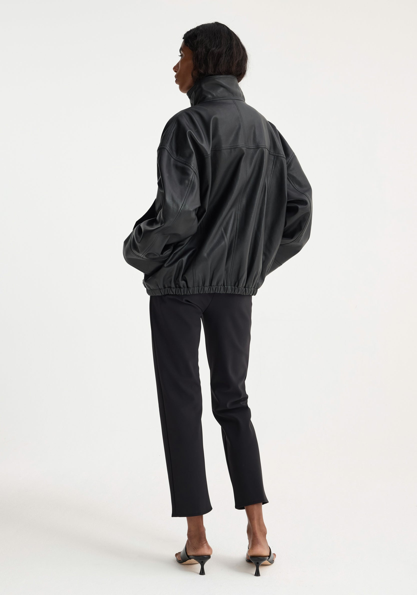 Back view of a person wearing a loose-fitting Reinvented leather jacket | black with a high collar, paired with black cropped pants and black heeled sandals.