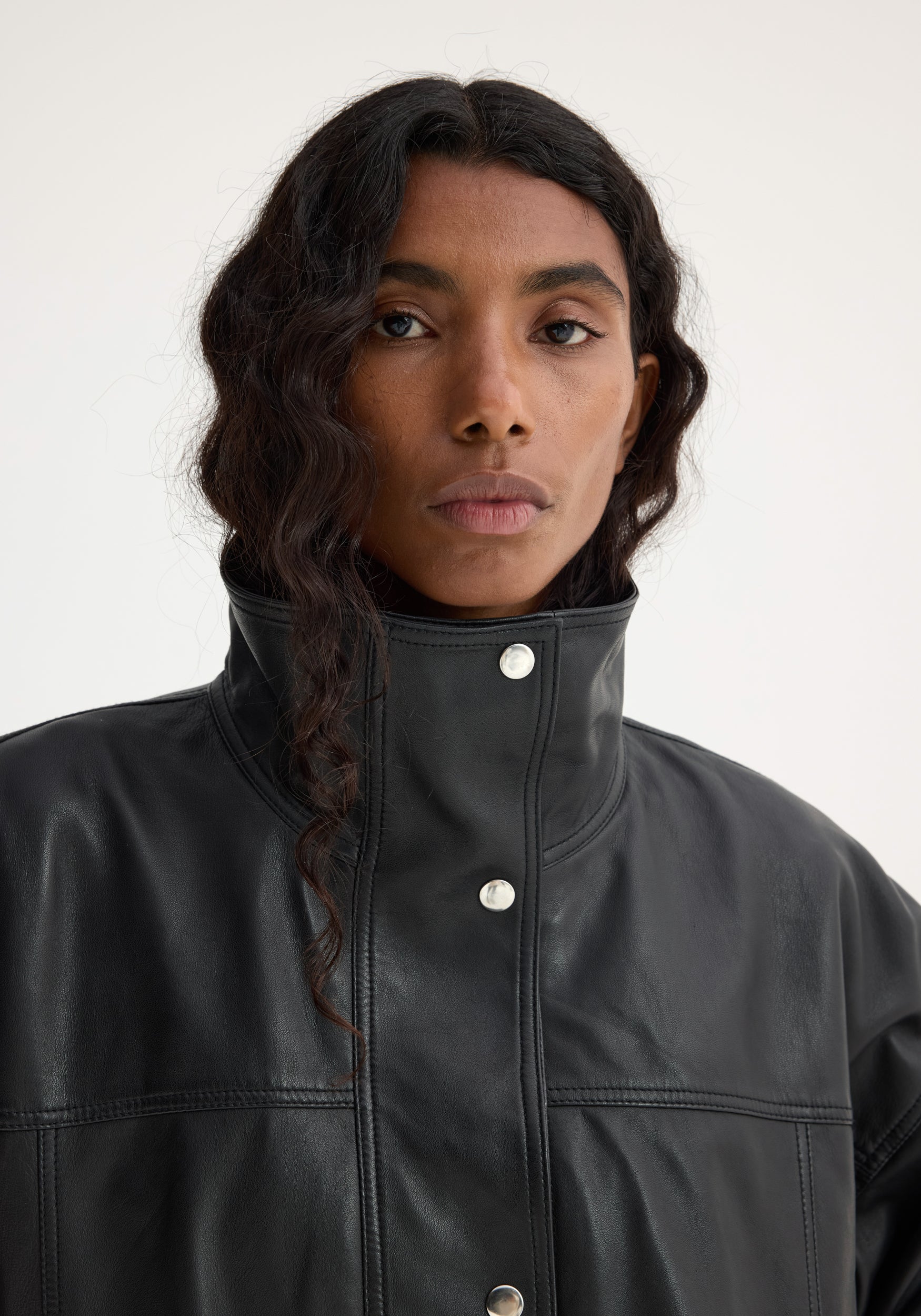 Close-up portrait of a woman wearing a high-collar black leather jacket with snap buttons, showcasing the textured material and sleek design of the Reinvented leather jacket | black.