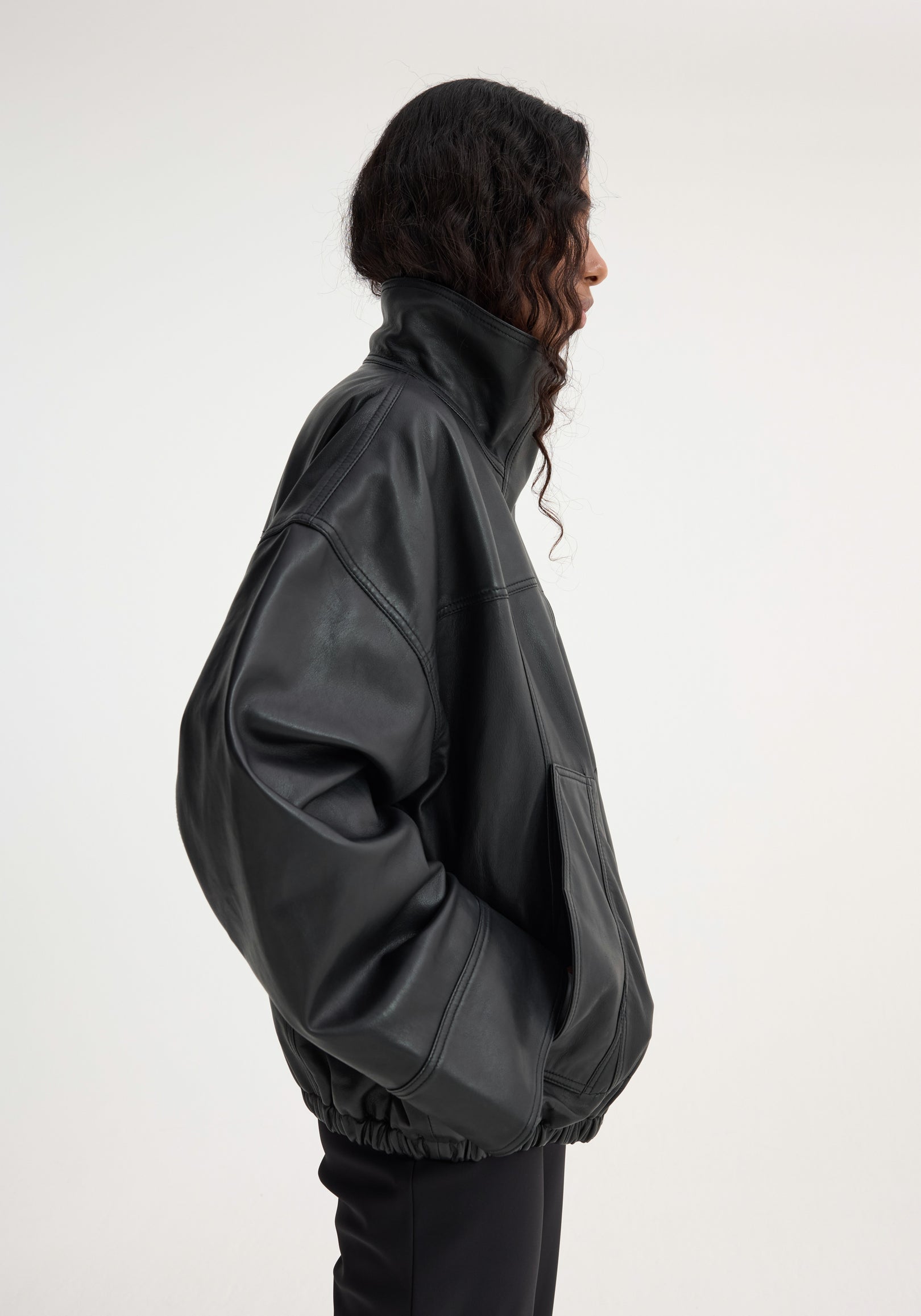 Profile view of a person wearing a black oversized reinvented leather jacket | black with a high collar and elasticated hem against a plain background.