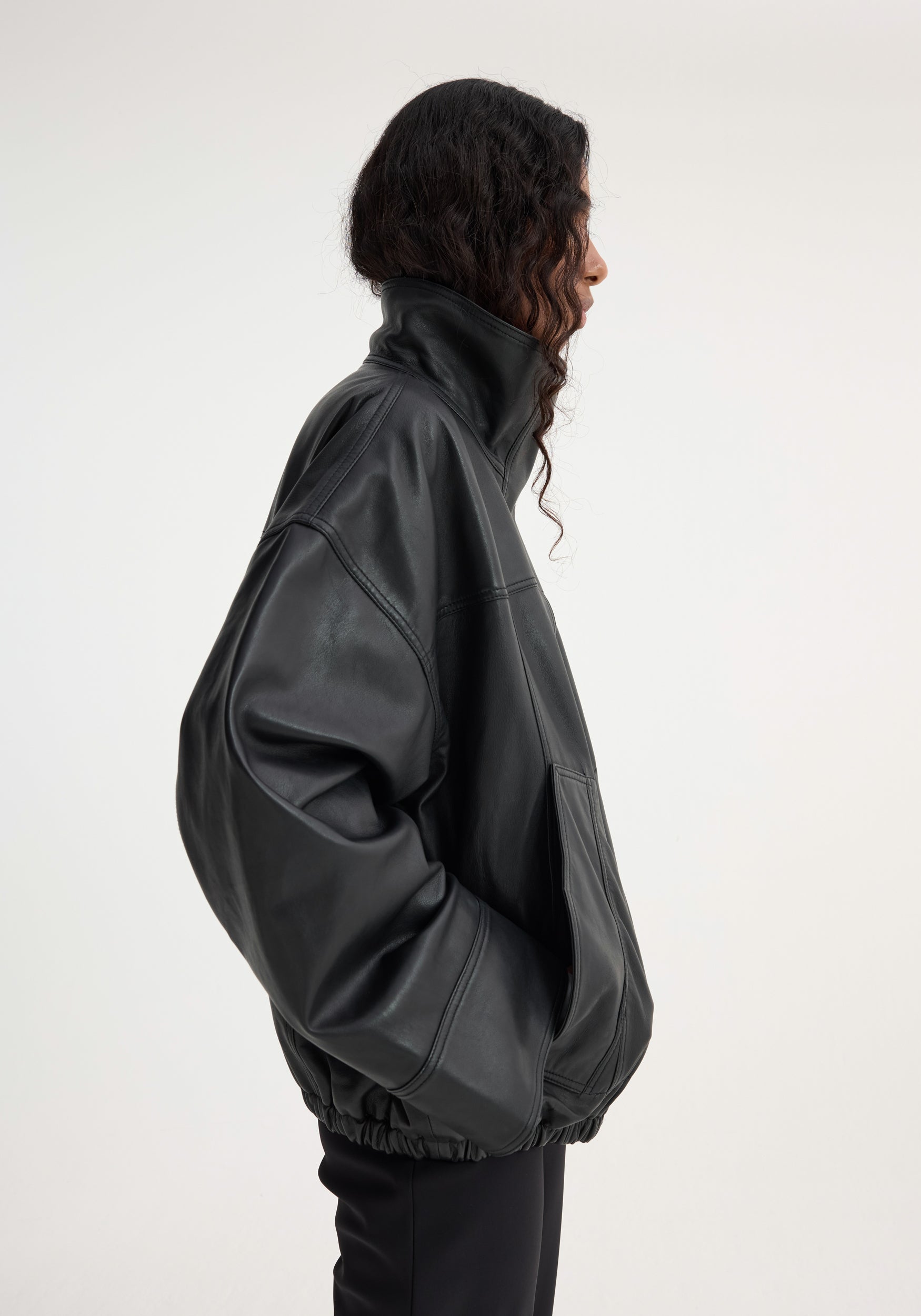 Profile view of a person wearing a black oversized reinvented leather jacket | black with a high collar and elasticated hem against a plain background.
