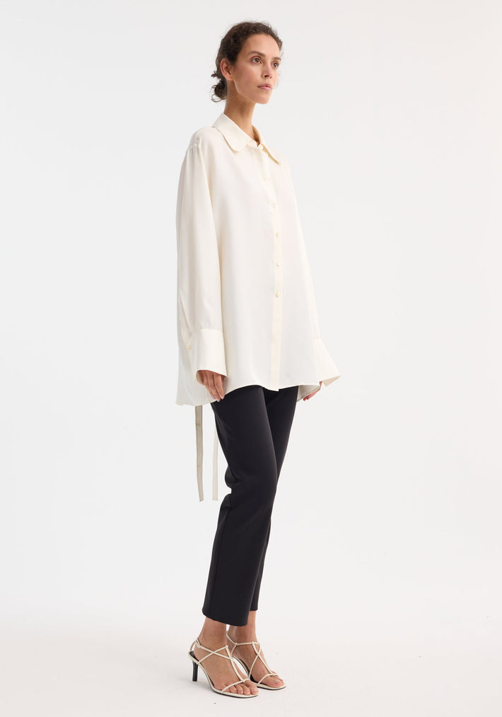 Model wearing Deconstructed silk shirt | cream, showcasing its oversized fit, exaggerated cuffs, open back with tie detail, paired with slim black trousers and neutral sandals.