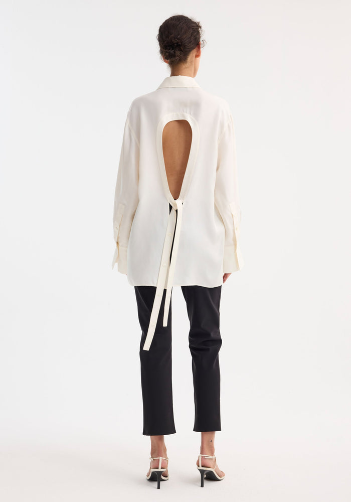 Back view of a woman in a Deconstructed silk shirt | cream, featuring an open back with teardrop cutout and fabric ties, paired with black trousers.