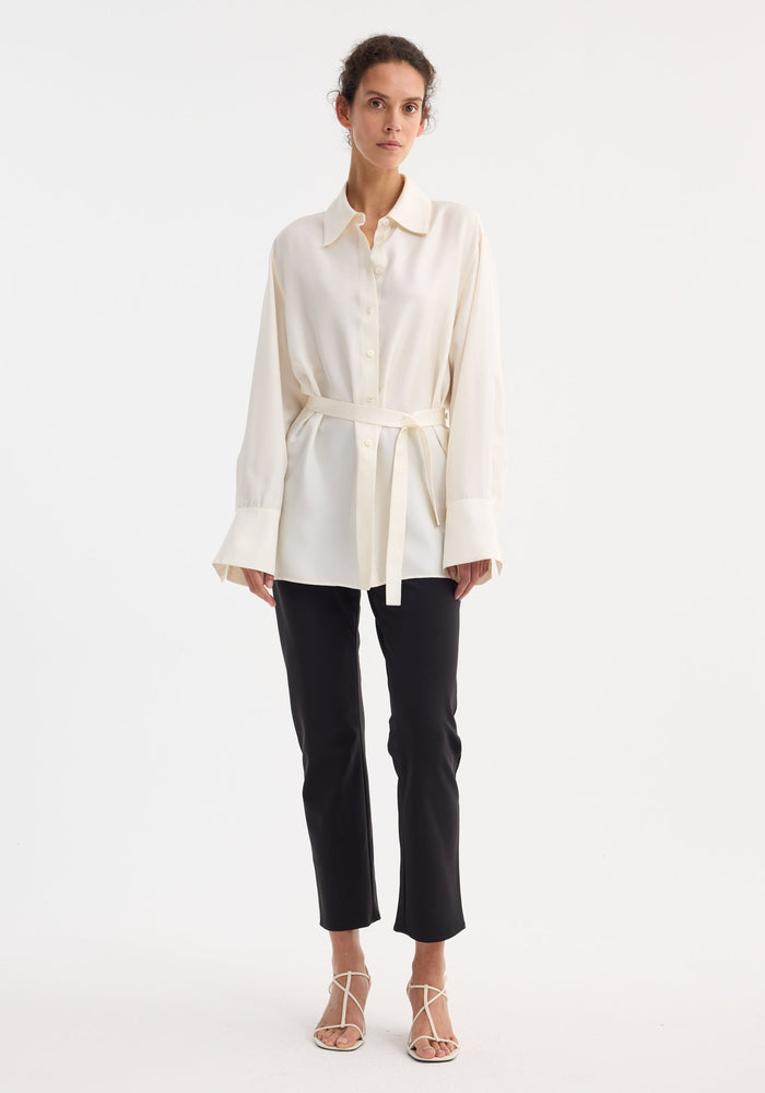 Model wearing Deconstructed silk shirt in cream, featuring a button-front, self-tie belt, and open back. Paired with slim black trousers and ivory sandals.