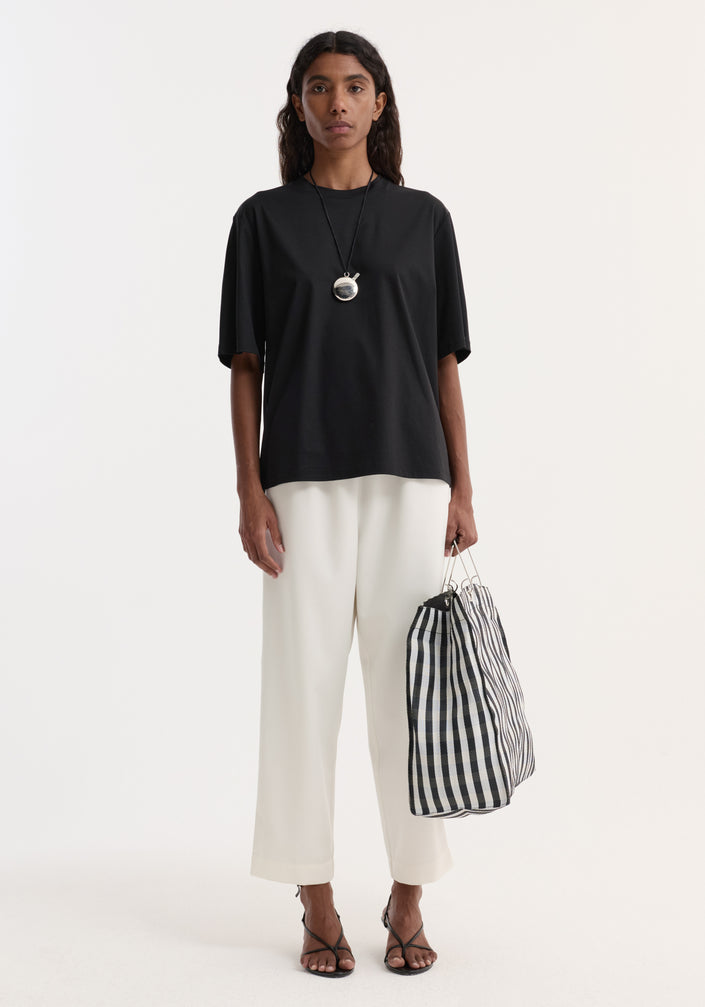 Person wearing an Oversized cotton t-shirt | black with wide-leg trousers, strappy sandals, and holding a striped tote bag, styled in a minimalist monochrome look.