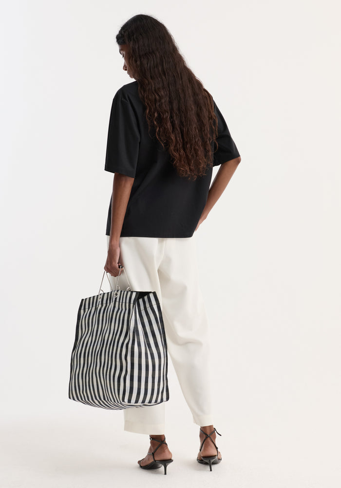 Back view of a woman in an Oversized cotton t-shirt, black, with wide-leg trousers, holding a striped tote bag, wearing strappy heels.