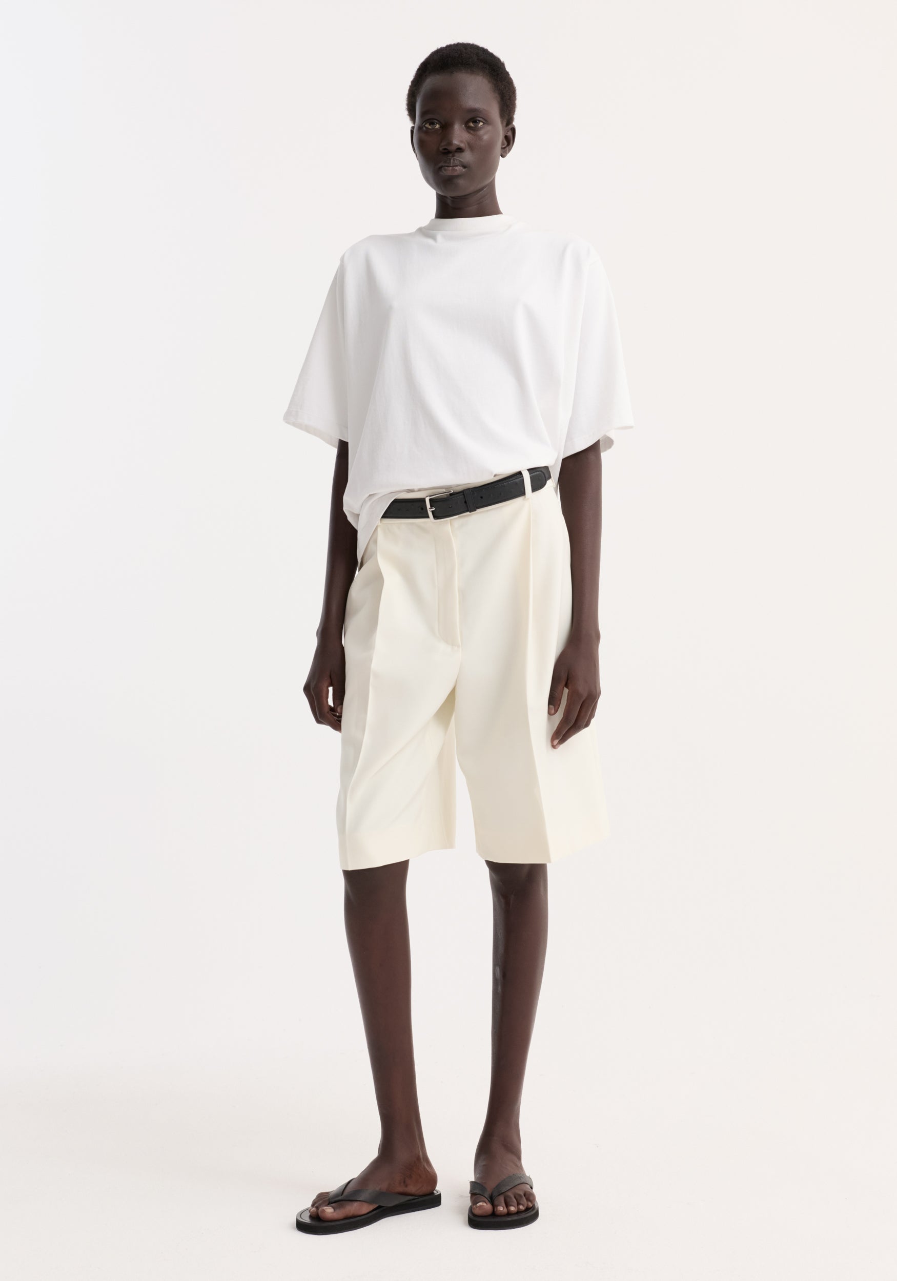 Model wearing an oversized cotton t-shirt | optic white with loose fit, paired with high-waisted cream shorts and black belt, styled with black sandals against a plain background.