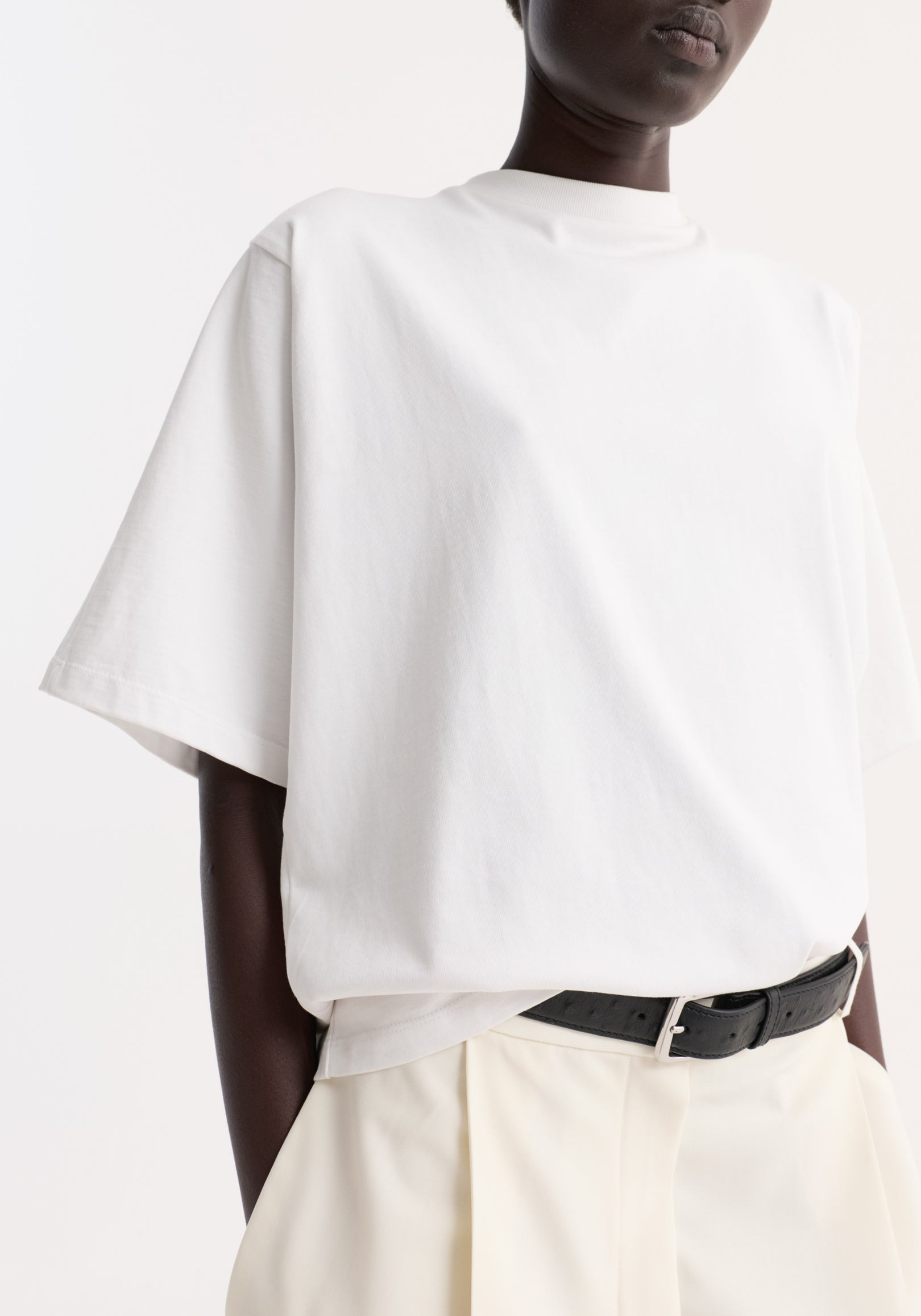 A close-up of a person wearing an oversized cotton t-shirt | optic white, styled with light-colored pants and a black belt, against a white background.