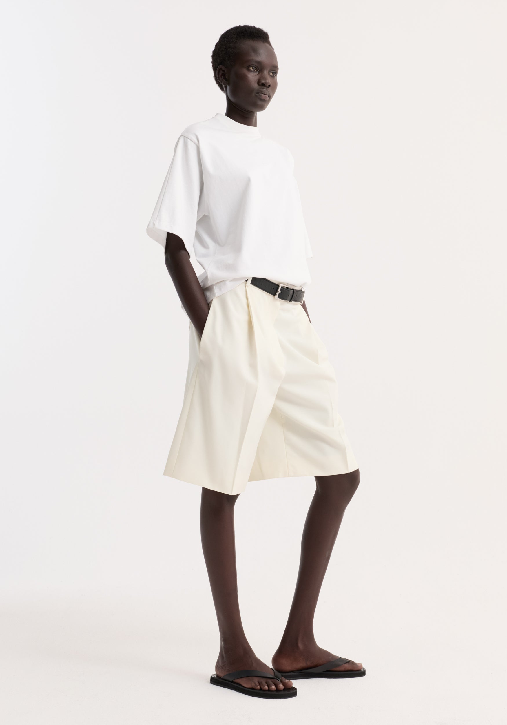 Model wearing an oversized cotton t-shirt | optic white with loose, light-colored shorts, black belt, and black sandals against a plain white background.