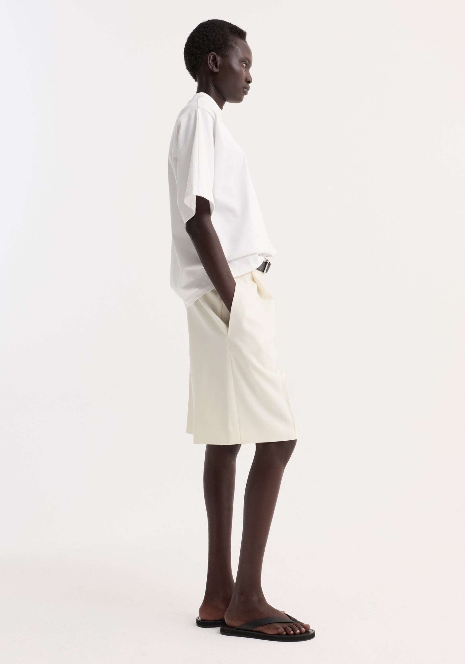 Side profile of a person wearing an oversized cotton t-shirt | optic white paired with light-colored shorts and black sandals against a plain white background.