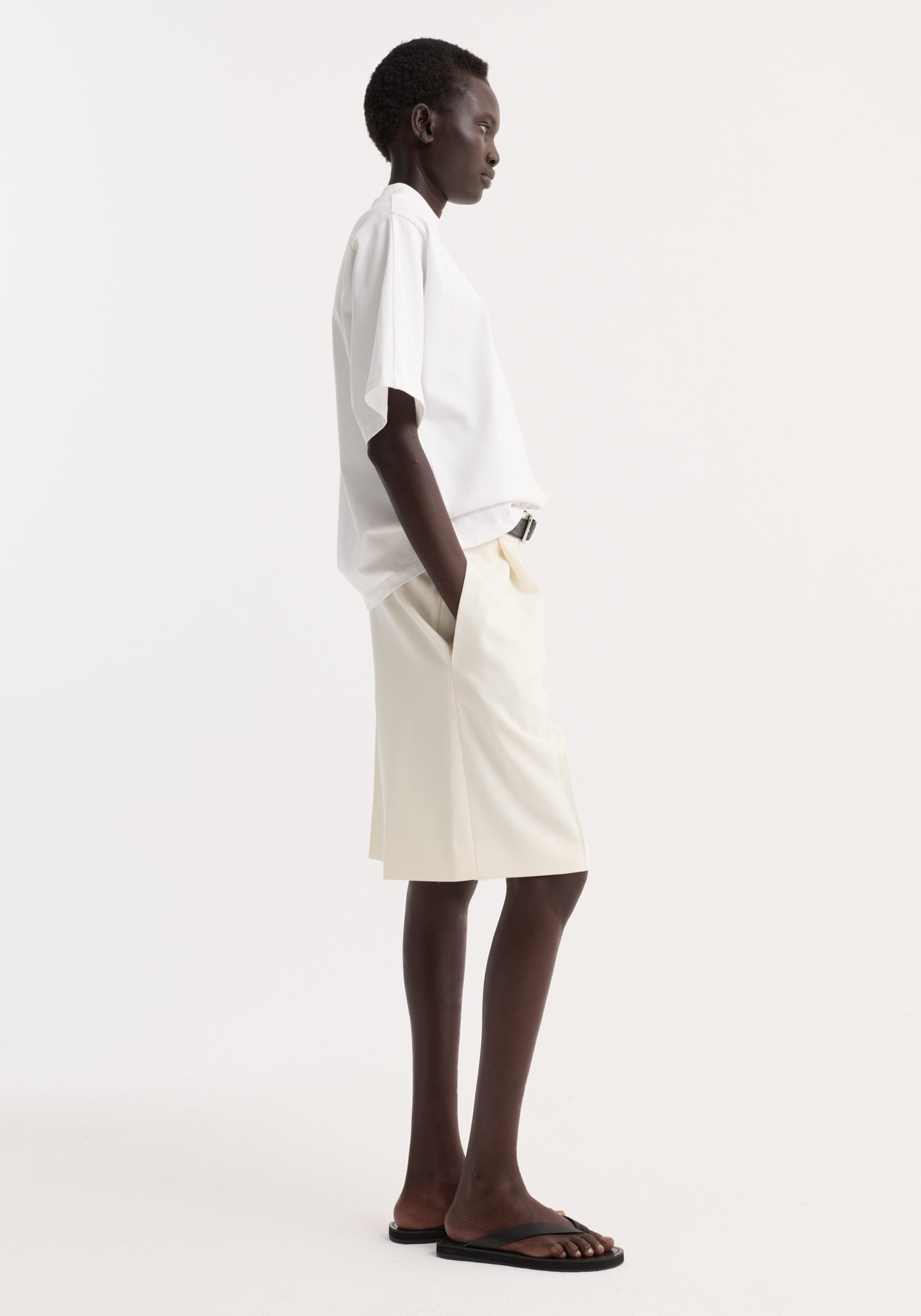 Side profile of a person wearing an oversized cotton t-shirt | optic white paired with light-colored shorts and black sandals against a plain white background.