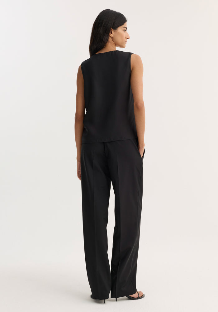 Woman wearing the Scoop-neck silk top in black, featuring a wide scoop neck and loose fit, paired with high-waisted trousers and heeled sandals.