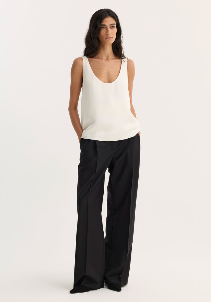 Woman wearing scoop-neck silk top | cream, tucked into high-waisted black trousers, hands in pockets, showcasing a minimalist, loose-fit design suitable for layering.