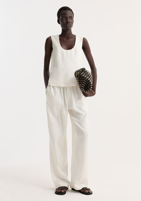 Tall woman models a scoop-neck silk top with a loose fit, paired with wide-leg trousers, holding a woven clutch. Simple, minimal, and elegant outfit.