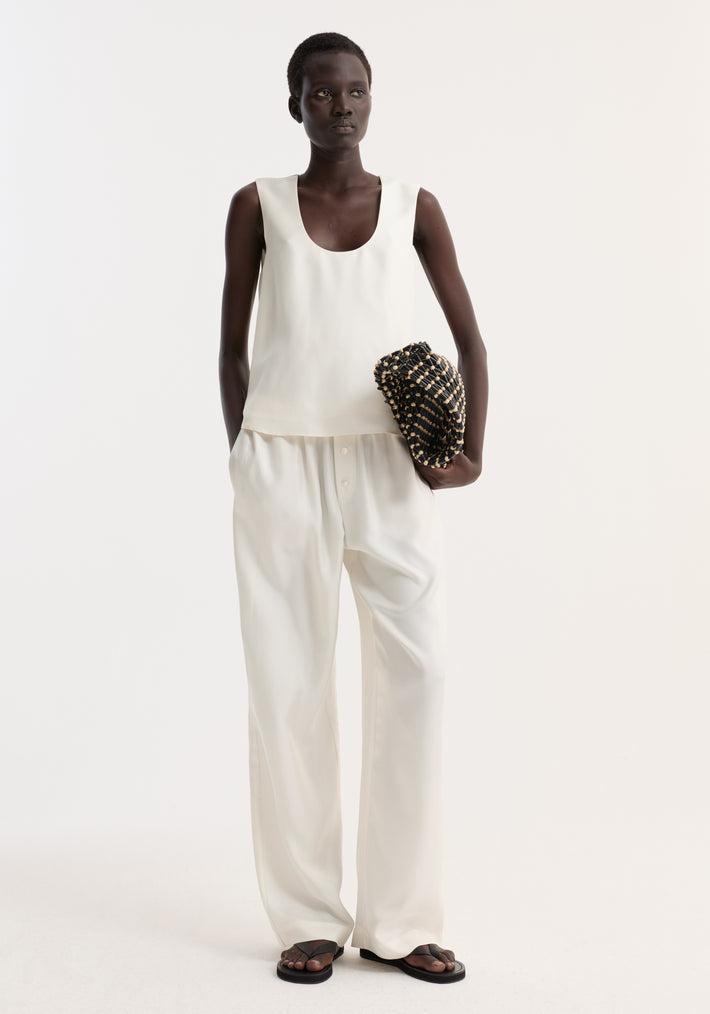 Tall woman models a scoop-neck silk top with a loose fit, paired with wide-leg trousers, holding a woven clutch. Simple, minimal, and elegant outfit.