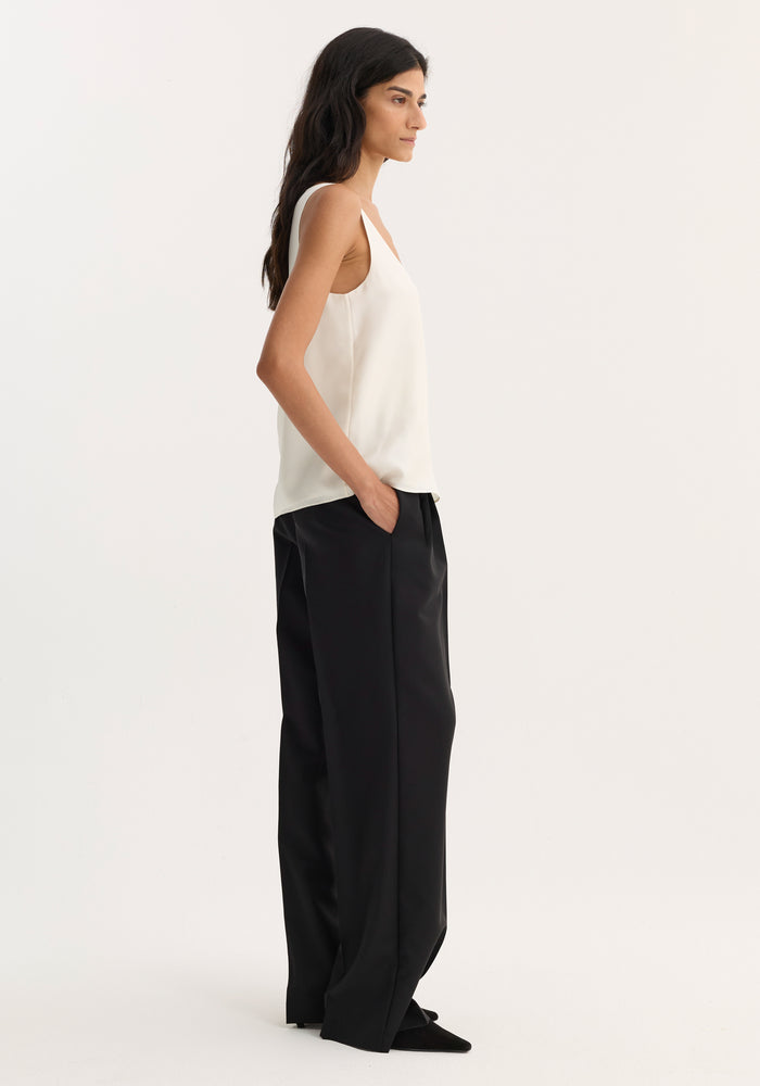 Model wearing a scoop-neck silk top in cream, featuring a loose fit and minimalist design, paired with high-waisted black trousers and pointed-toe shoes.