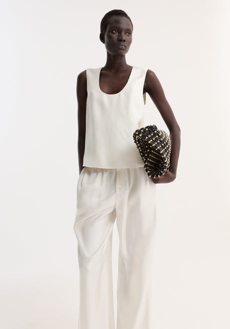 Tall woman models a scoop-neck silk top with a loose fit, paired with wide-leg trousers, holding a woven clutch. Simple, minimal, and elegant outfit.