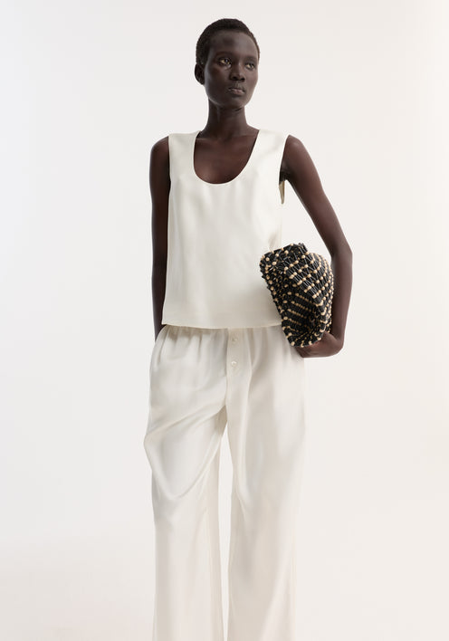 Tall woman models a scoop-neck silk top with a loose fit, paired with wide-leg trousers, holding a woven clutch. Simple, minimal, and elegant outfit.