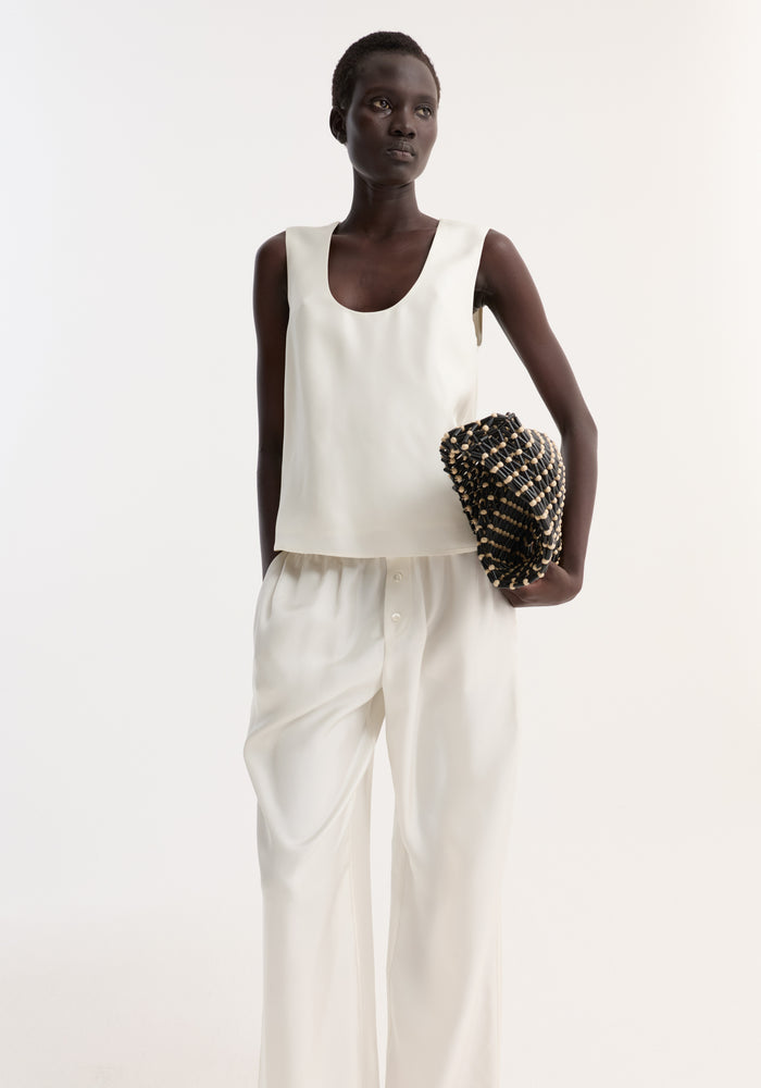 Woman wearing Scoop-neck silk top | cream, showcasing a loose fit and scoop neck, paired with wide-leg trousers. Holding a beaded clutch.