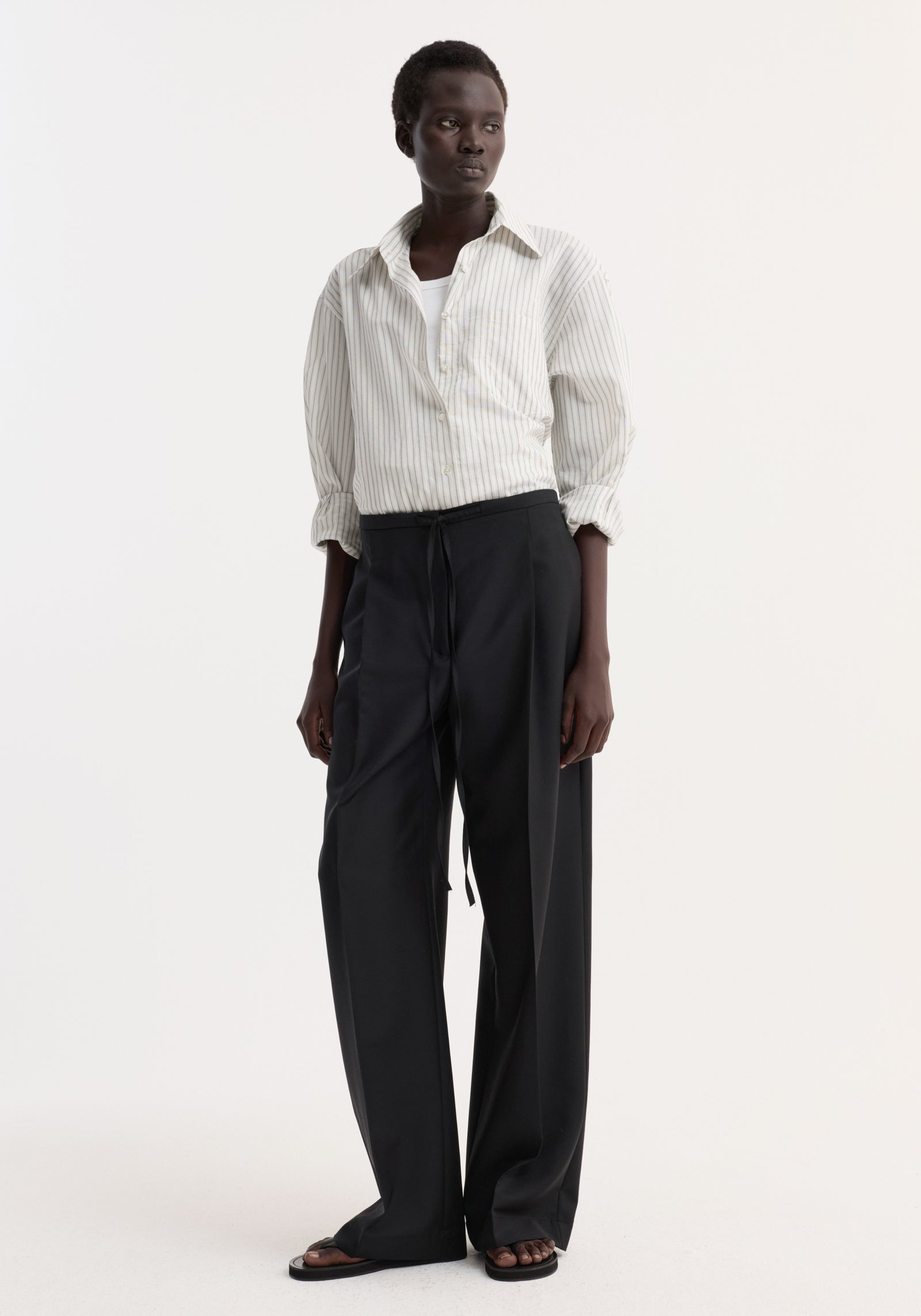 Model wearing Relaxed drawstring trousers in black, featuring a long, wide-leg silhouette with a drawstring waistband and front pleats, paired with a white shirt.