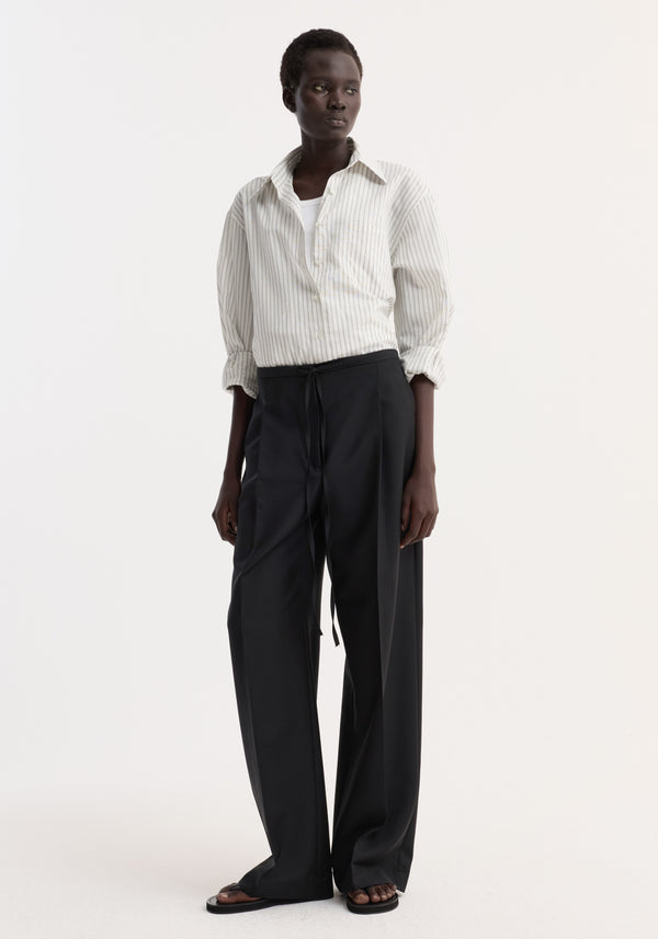 Model wearing Relaxed drawstring trousers in black, featuring a long, wide-leg silhouette with a drawstring waistband and front pleats, paired with a white shirt.