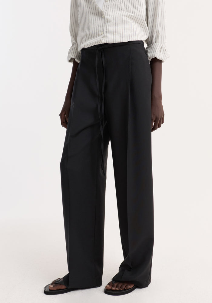 Model wearing Relaxed drawstring trousers | black, featuring wide legs, slim tie waistband, front pleats, and back welt pockets, paired with flat sandals.