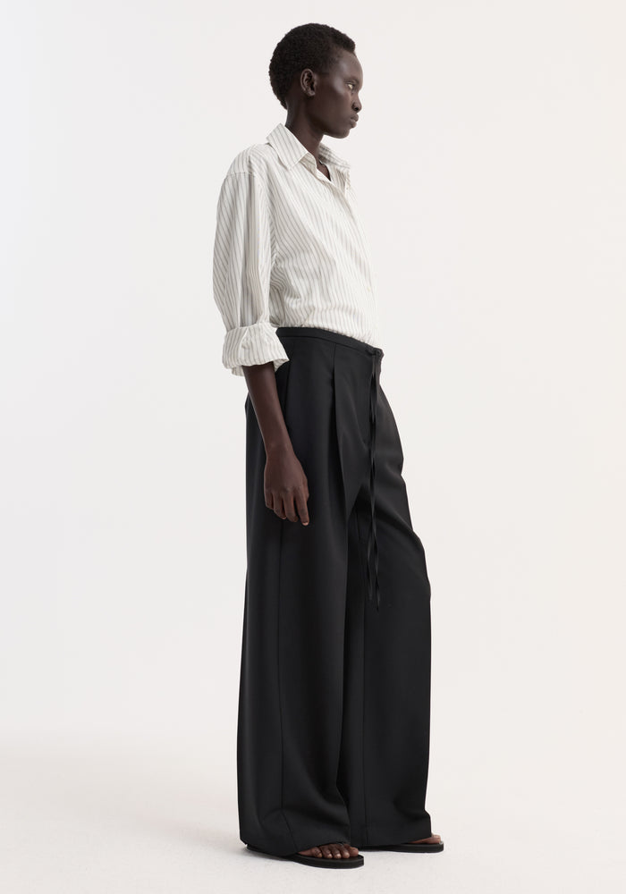 Profile view of a person wearing Relaxed drawstring trousers in black, featuring a long, wide-leg design with a drawstring waistband, styled with a button-up shirt.