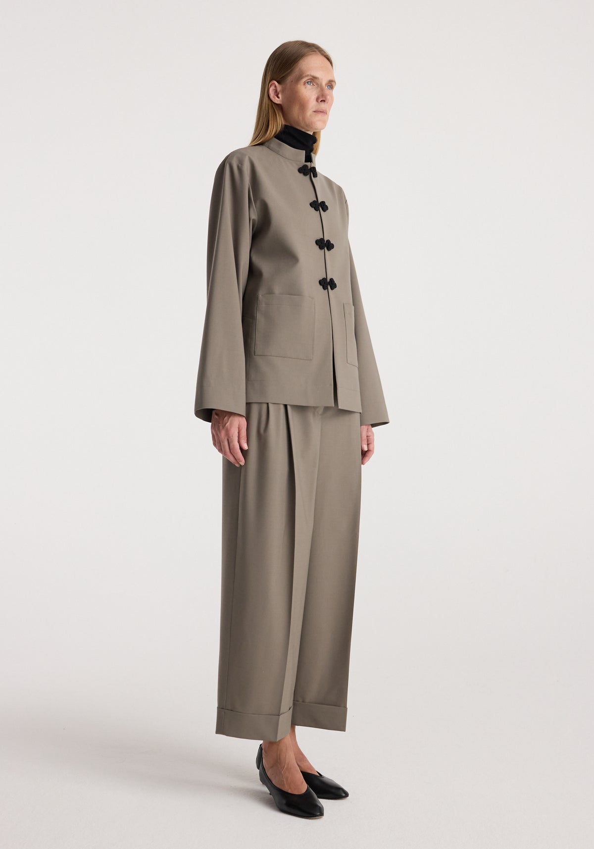 Woman wearing high-waist turn-up trousers | taupe paired with a matching jacket and black shoes against a plain background.