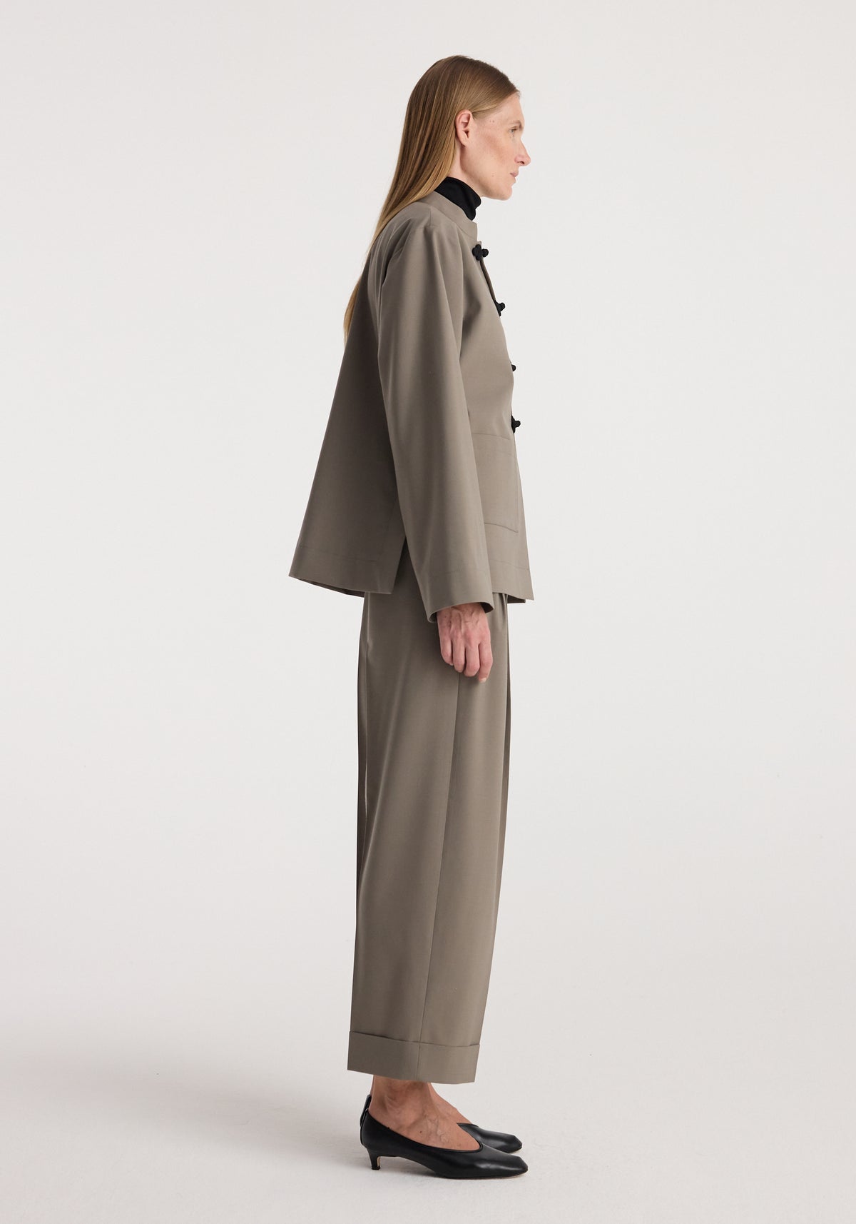 Side view of a woman wearing high-waist turn-up trousers | taupe styled with a matching loose-fitting jacket and black low-heeled shoes against a plain background.