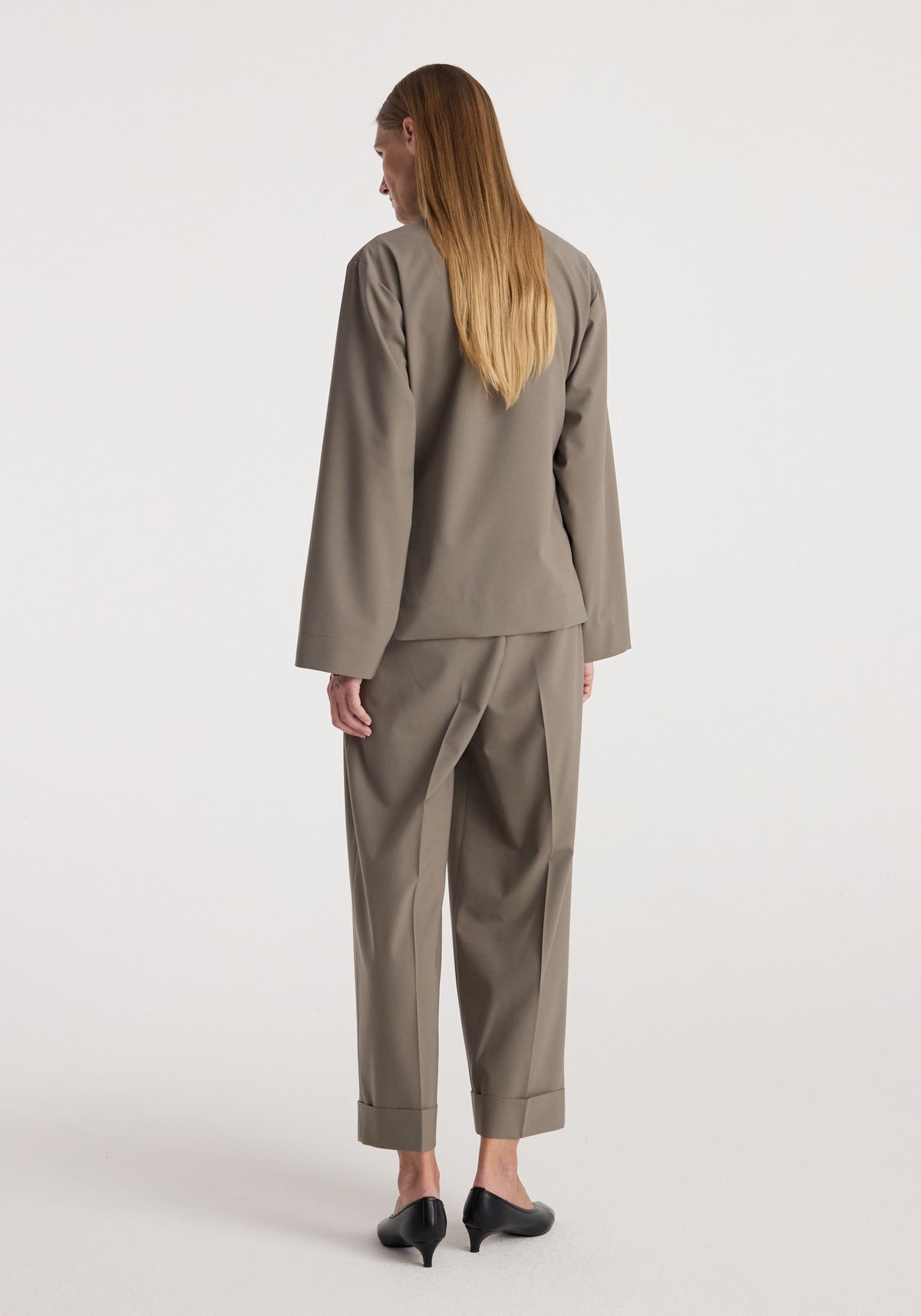 Back view of a woman wearing high-waist turn-up trousers | taupe paired with a matching long-sleeve top and black heeled shoes against a plain white background.