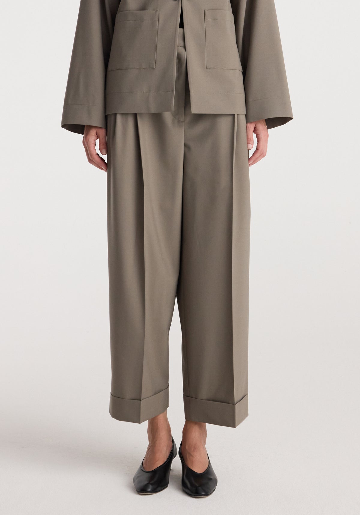 Front view of a person wearing taupe high-waist turn-up trousers | taupe, paired with a matching jacket and black low-heeled shoes against a plain background.