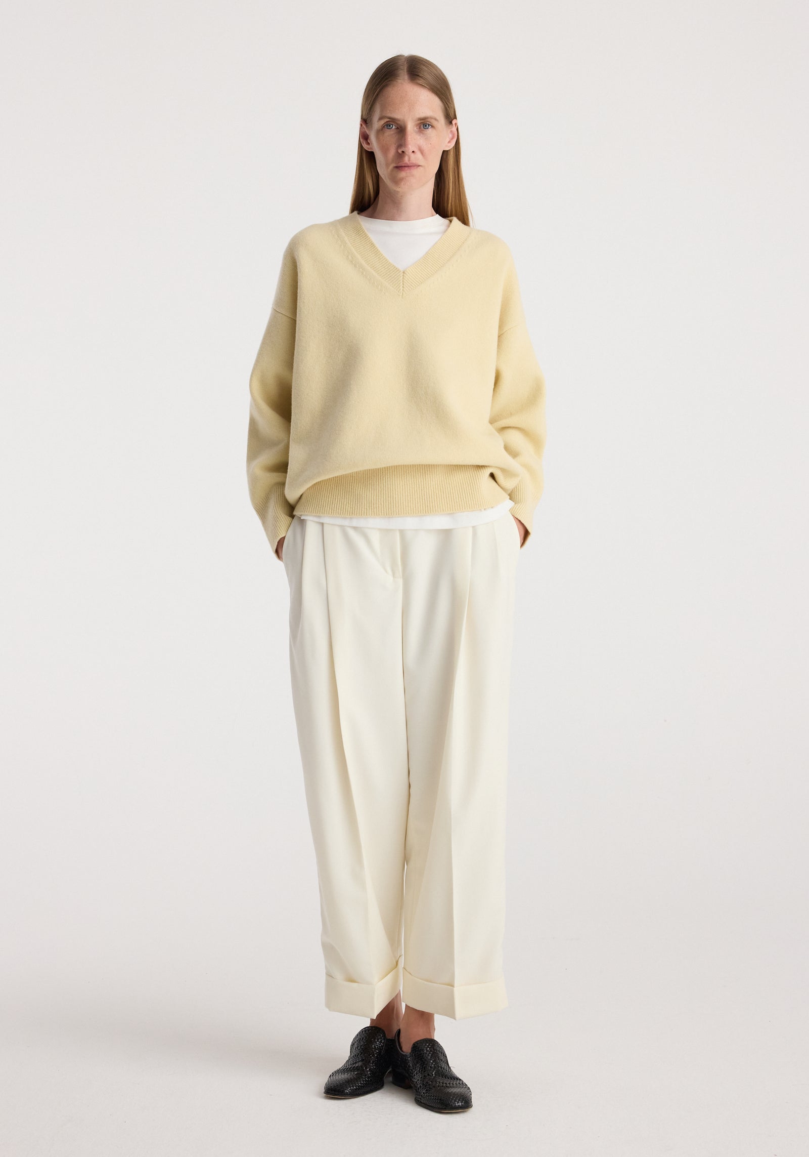 Woman wearing high-waist turn up trousers | off white paired with a loose beige sweater and black shoes, standing against a plain light background.