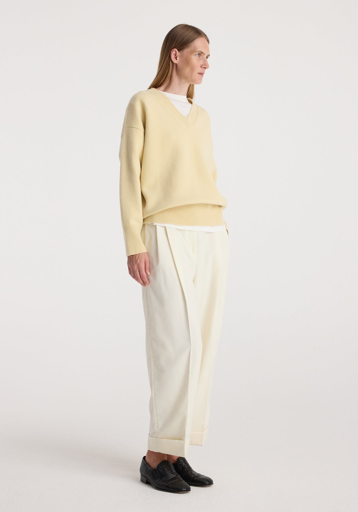 Woman wearing off white High-waist turn up trousers paired with a light beige oversized V-neck sweater and black woven loafers against a plain background.