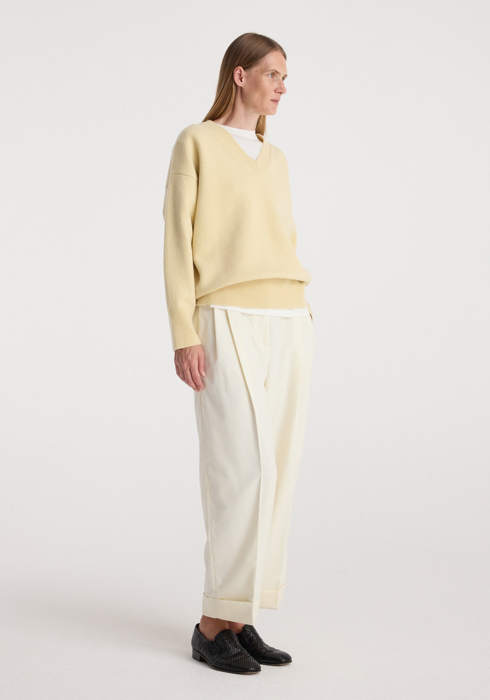 Woman wearing off white High-waist turn up trousers paired with a light beige oversized V-neck sweater and black woven loafers against a plain background.