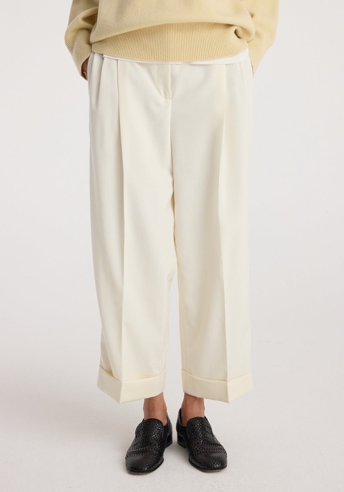 Front view of a person wearing High-waist turn up trousers | off white with wide cuffs and pleats, paired with a beige sweater and black woven shoes on a plain background.