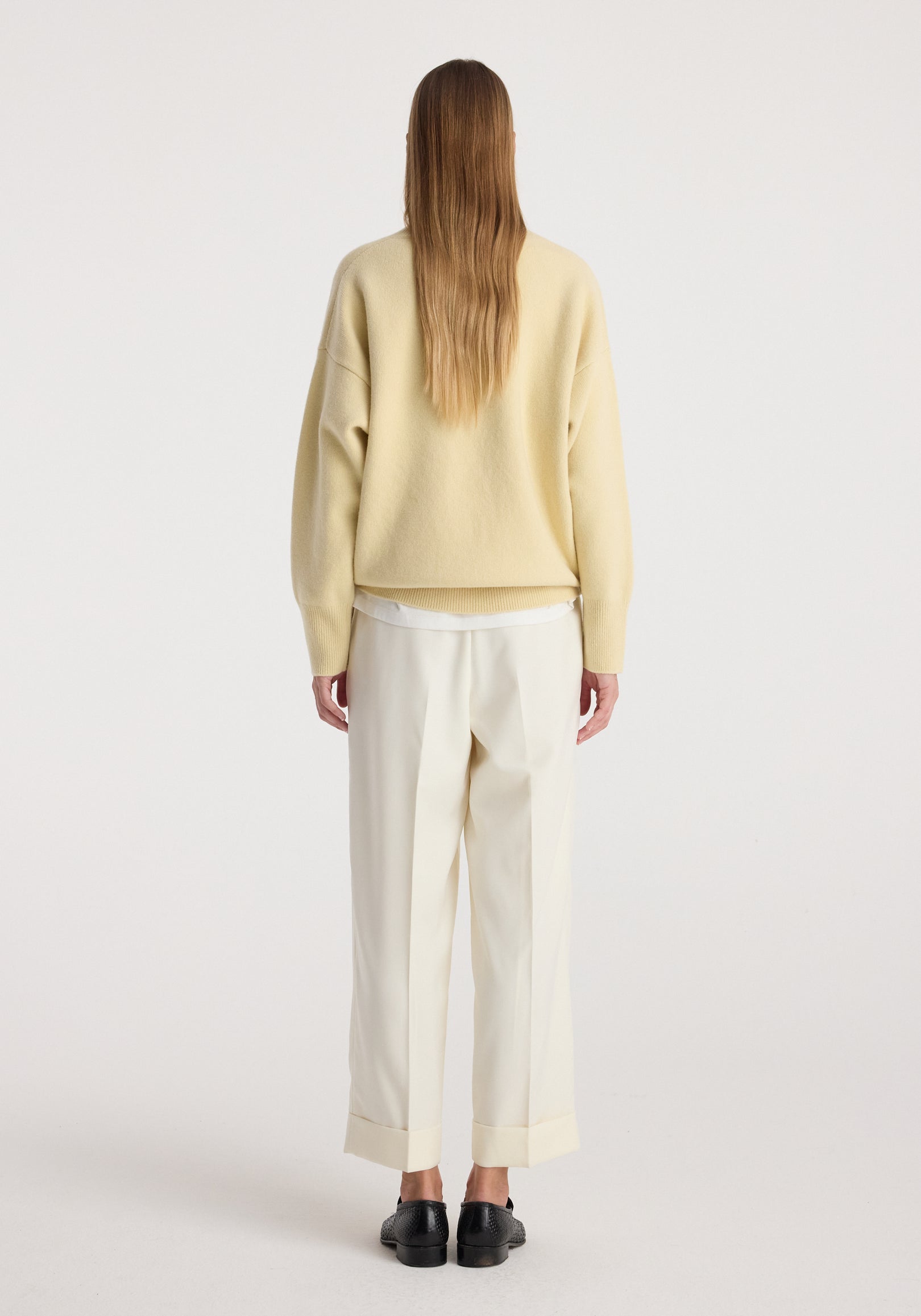 Back view of a person wearing High-waist turn up trousers | off white paired with a beige sweater and black shoes on a plain background.
