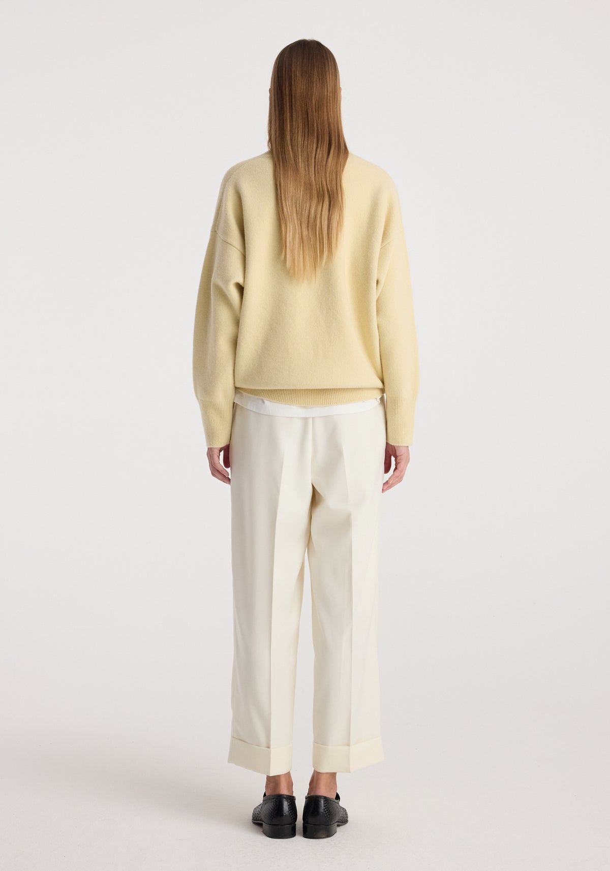 Back view of a person wearing High-waist turn up trousers | off white paired with a beige sweater and black shoes on a plain background.
